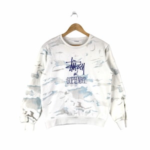 May include: White long-sleeve sweatshirt with a blue and white camouflage pattern. The sweatshirt has a blue embroidered logo that says "Stussy and Sophnet."