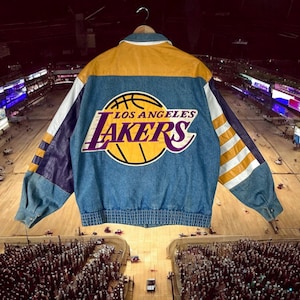 Puede incluir: Una chaqueta vaquera vintage de Los Angeles Lakers. La chaqueta presenta un cuerpo de mezclilla azul con cuello y panel trasero de cuero amarillo. El logotipo de los Lakers se muestra en la espalda. Mangas con detalles morados y blancos.