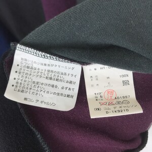 Puede incluir: Un primer plano de una etiqueta de tela negra y morada con texto blanco que detalla las instrucciones de cuidado de la prenda. La etiqueta incluye el nombre de la marca "Commes des Garcons" y el n&uacute;mero de producto "D-TK9210".