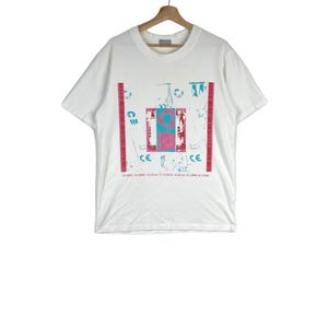 May include: White t-shirt with a graphic print of a colorful abstract design. The design includes geometric shapes, lines, and the letters "CE". The text "BILLION BILLION BILLION BILLION BILLION BILLION" is printed at the bottom of the design.