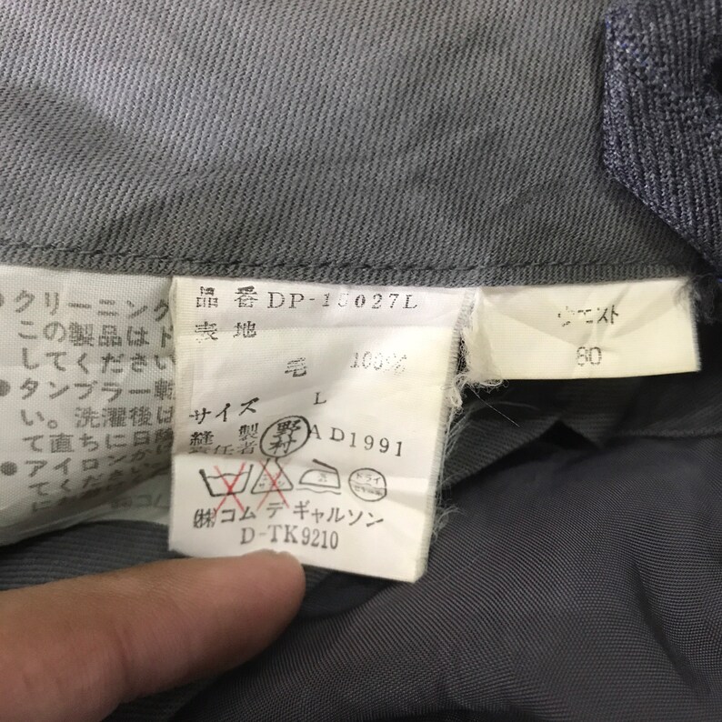 May include: Close-up of a gray garment tag with Japanese text and care symbols. The tag includes the product code DP-10027L, size L, and waist measurement of 80. The fabric composition is 100% wool. The brand name is also visible.