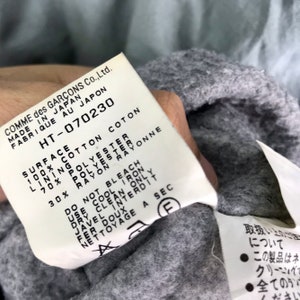 Op de afbeelding: Een wit label met zwarte tekst met details over de stofsamenstelling en de wasinstructies voor een kledingstuk. Het label luidt "COMME des GAR&Ccedil;ONS Co., Ltd. MADE IN JAPAN FABRIQUE AU JAPON HT-070230 SURFACE 100% COTTON COTON LINING 70% POLYESTER 30% RAYON DO NOT BLEACH USE COOL IRON DRY CLEAN ONLY JAVEL INTERDIT FER DOUX NETTOYAGE A SEC"
