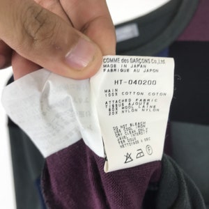 Puede incluir: Una etiqueta blanca con texto negro que detalla la composici&oacute;n y las instrucciones de cuidado de una prenda. La etiqueta dice "COMME des GARCONS Co., Ltd. MADE IN JAPAN FABRIQUE AU JAPON HT-040200 MAIN 100% COTTON COTON ATTACHED FABRIC TISSUS AJOUTE 80% WOOL LATNE 20% NYLON NYLON DO NOT BLEACH USE COOL IRON DRY CLEAN ONLY JAVEL INTERDTT FER DOUX NETTOYAGE A SEC"