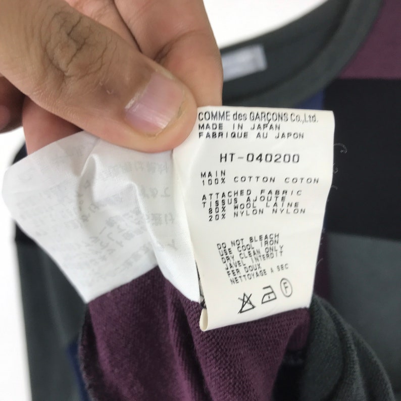 Puede incluir: Una etiqueta blanca con texto negro que detalla la composici&oacute;n y las instrucciones de cuidado de una prenda. La etiqueta dice "COMME des GARCONS Co., Ltd. MADE IN JAPAN FABRIQUE AU JAPON HT-040200 MAIN 100% COTTON COTON ATTACHED FABRIC TISSUS AJOUTE 80% WOOL LATNE 20% NYLON NYLON DO NOT BLEACH USE COOL IRON DRY CLEAN ONLY JAVEL INTERDTT FER DOUX NETTOYAGE A SEC"