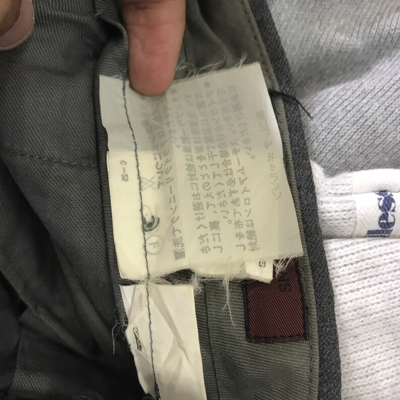 May include: Close-up of a gray pair of pants with a white tag attached. The tag has Japanese text and a small logo. A portion of a gray sweatshirt is visible in the background.