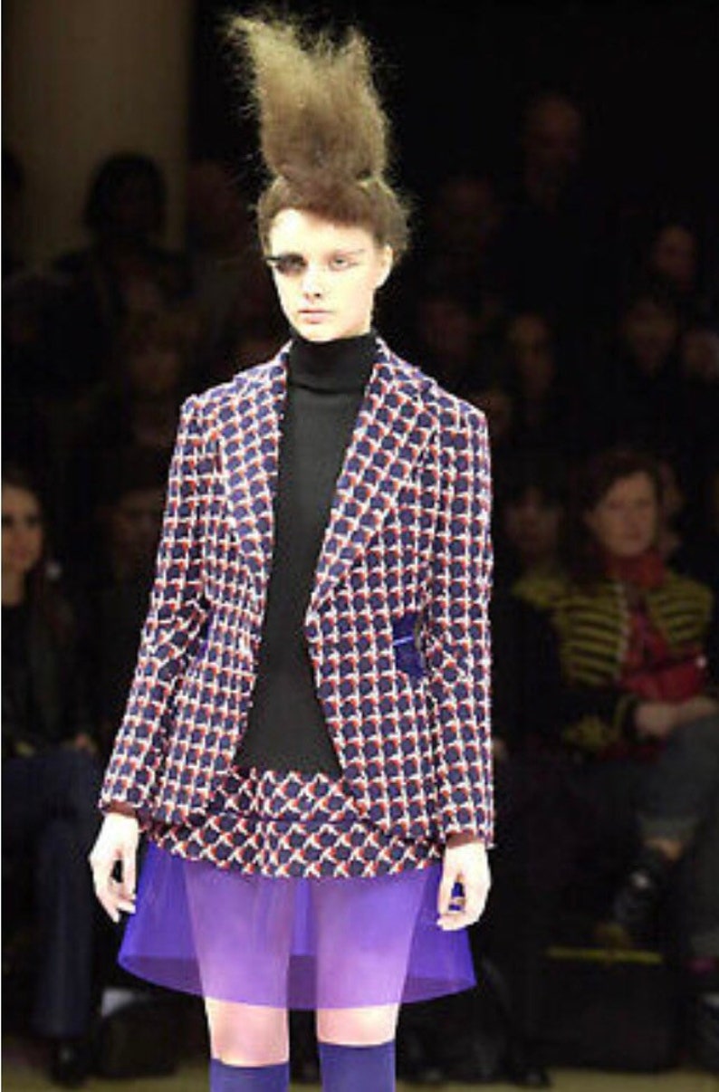 May include: A model in a fashion show wearing a patterned blazer, matching skirt, and purple sheer skirt over purple shorts. The blazer and skirt have a blue and red geometric design. The model is also wearing a black turtleneck.