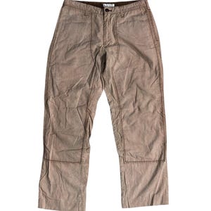 Puede incluir: Pantalones color beige con costuras visibles. Los pantalones tienen cierre de botón y cremallera, con refuerzos en las rodillas. Las costuras son de color marrón oscuro, creando contraste. Los pantalones son de material suave.