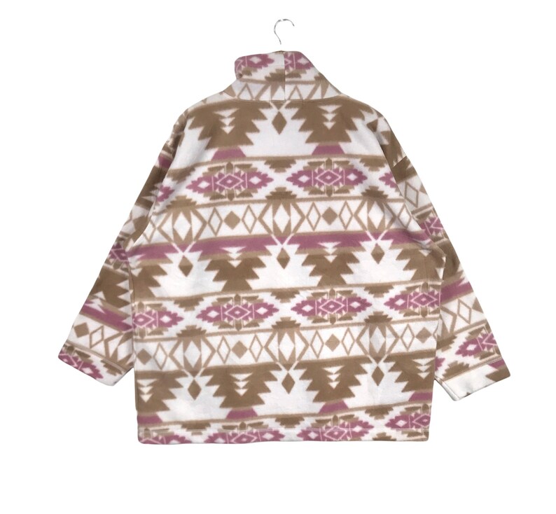 Puede incluir: Una chaqueta de forro polar blanca con un patr&oacute;n geom&eacute;trico marr&oacute;n y rosa. La chaqueta tiene un cuello alto y una cremallera.
