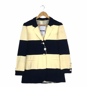 May include: A striped blazer with a white and navy blue color scheme. The blazer has a single button closure and a label that reads "Moschino Couture".