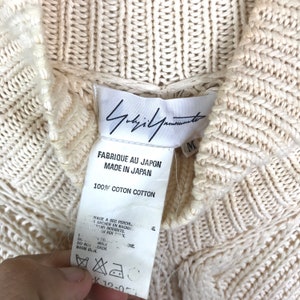 May include: A white cotton sweater with a cable knit pattern. The sweater has a high neck and a label that reads "Yohji Yamamoto" and "Fabrique au Japon Made in Japan 100% Coton Cotton".