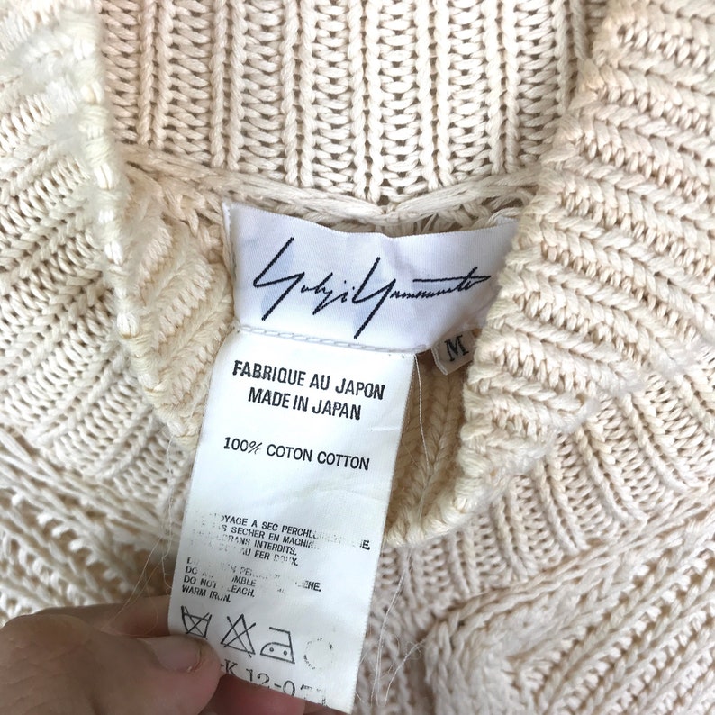 May include: A white cotton sweater with a cable knit pattern. The sweater has a high neck and a label that reads "Yohji Yamamoto" and "Fabrique au Japon Made in Japan 100% Coton Cotton".