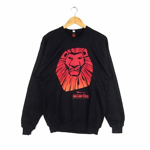 Peut inclure: Sweat-shirt noir avec un lion rouge et orange et le texte "Disney Presents The Lion King The Broadway Musical".