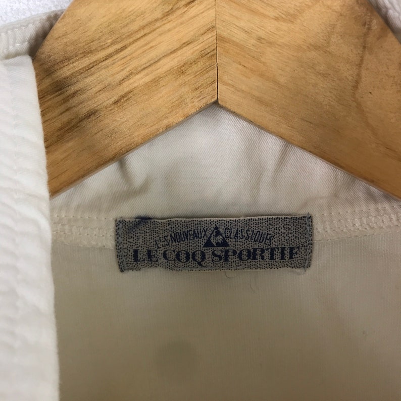 May include: White shirt with a gray label that reads "Le Coq Sportif" with a blue triangle logo.