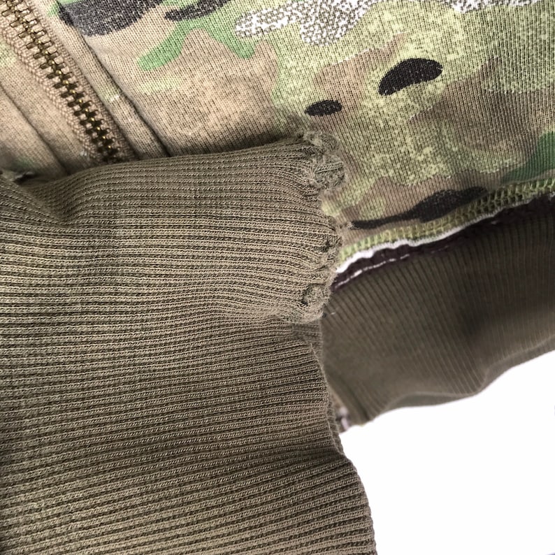 May include: Close-up of a green and brown camouflage fabric with a ribbed fabric underneath. The ribbed fabric has a frayed edge.