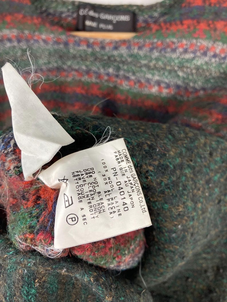 May include: A close-up of a black and white label with the text "COMME des GAR&Ccedil;ONS Co., Ltd. MADE IN JAPAN FABRIQUE AU JAPON PN-040140 ALBASA 100% WOOL LAINE 30% AL DO NOT BLEACH COOL IRON DRY CLEAN ONLY JAVEL INTERDIT FER DOUX NETTOYAGE A SEC"