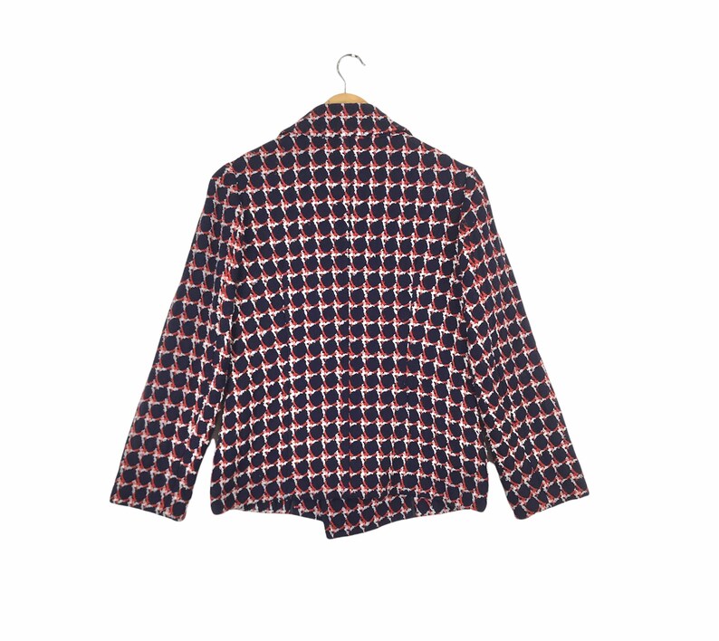 May include: A navy blue blazer with a repeating pattern of red and white shapes. The blazer is hanging on a wooden hanger against a white background. The blazer has long sleeves and a classic collar.