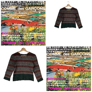 May include: A striped sweater in shades of green, red, and gray. The sweater has a crew neck and long sleeves. The sweater is hanging on a wooden hanger. The text "COMME des GARCONS" is visible in the image.