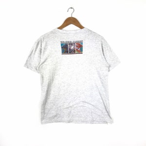 May include: A grey t-shirt with a graphic print on the back. The graphic features three characters from the anime series Neon Genesis Evangelion. The text "Neon Genesis Evangelion" is printed above the graphic.