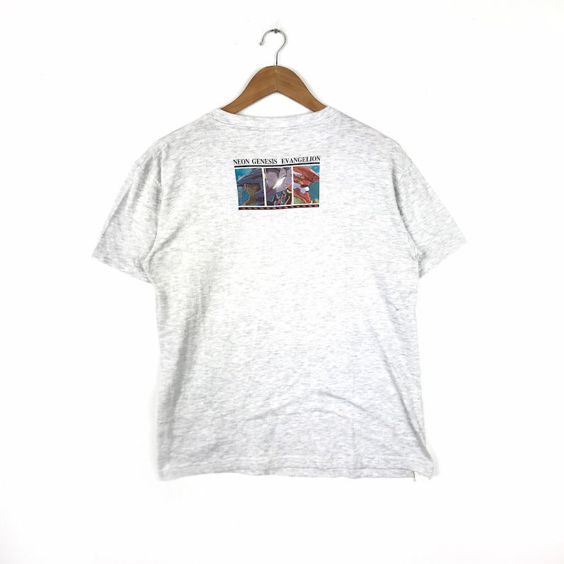 May include: A grey t-shirt with a graphic print on the back. The graphic features three characters from the anime series Neon Genesis Evangelion. The text "Neon Genesis Evangelion" is printed above the graphic.