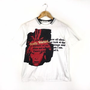 Può includere: Maglietta bianca con una stampa grafica in nero e rosso del viso di Andy Warhol. La maglietta ha un colletto a righe bianche e nere e il testo "If you want to know all about Andy Warhol just look at the surface: of my paintings and films and me and there I am. There's nothing and it."