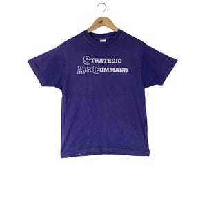 May include: A purple t-shirt with the text "STRATEGIC AIR COMMAND" in white block letters. The shirt is hanging on a wooden hanger against a white background. The t-shirt has short sleeves.
