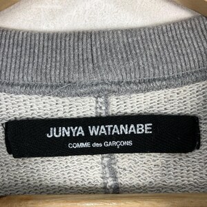 以下が含まれることがあります： グレーのスウェットシャツの襟のクローズアップ。黒いラベルには、白い文字で「JUNYA WATANABE」と「COMME des GAR&Ccedil;ONS」と書かれています。スウェットシャツは木製のハンガーに吊るされています。