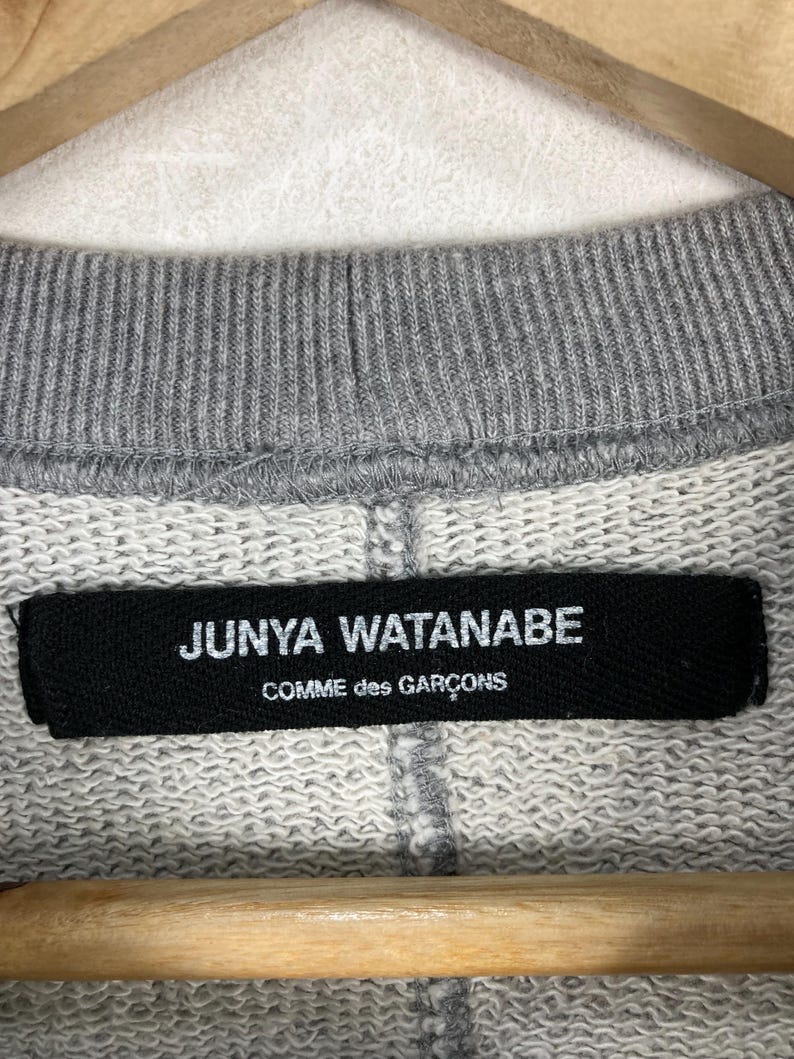 以下が含まれることがあります： グレーのスウェットシャツの襟のクローズアップ。黒いラベルには、白い文字で「JUNYA WATANABE」と「COMME des GAR&Ccedil;ONS」と書かれています。スウェットシャツは木製のハンガーに吊るされています。