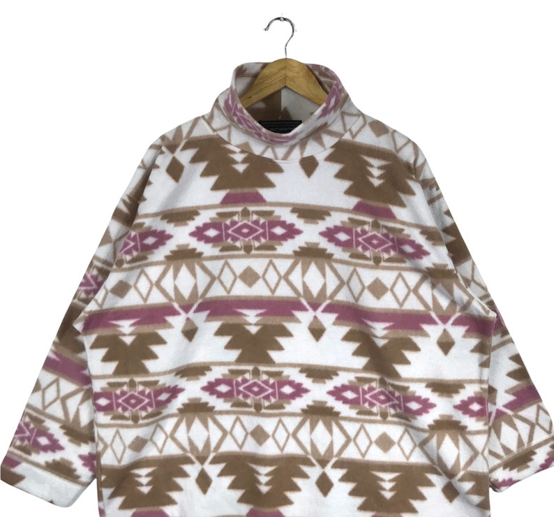 Puede incluir: Sudadera de vell&oacute;n blanca con un patr&oacute;n geom&eacute;trico marr&oacute;n y rosa. La sudadera tiene cuello alto y mangas largas.
