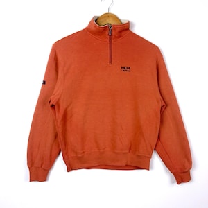 Puede incluir: Una sudadera naranja quemada de media cremallera con un logotipo MCM negro y el texto "Legere" bordado en la parte delantera.