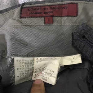 May include: Close-up of the inside waistband of a pair of gray denim pants. The label reads "COMME des GAR&Ccedil;ONS HOMME DEUX" and includes size and care instructions. The waist size is 80.
