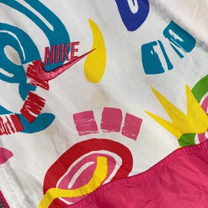 K&ouml;nnte beinhalten: Wei&szlig;e Nike Windjacke mit einem lebendigen abstrakten Muster in Rot, Blau, Gelb und Gr&uuml;n. Das Nike-Logo ist rot gestickt. Der untere Teil der Jacke ist leuchtend pink.