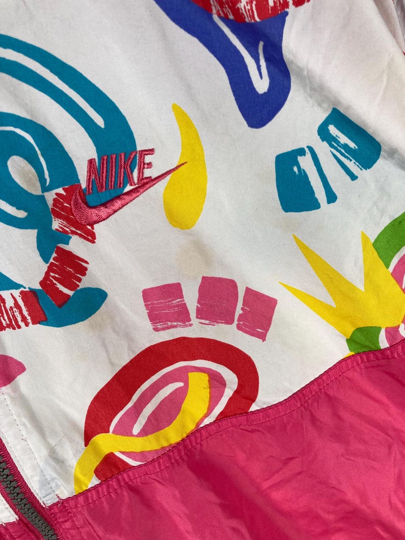 K&ouml;nnte beinhalten: Wei&szlig;e Nike Windjacke mit einem lebendigen abstrakten Muster in Rot, Blau, Gelb und Gr&uuml;n. Das Nike-Logo ist rot gestickt. Der untere Teil der Jacke ist leuchtend pink.