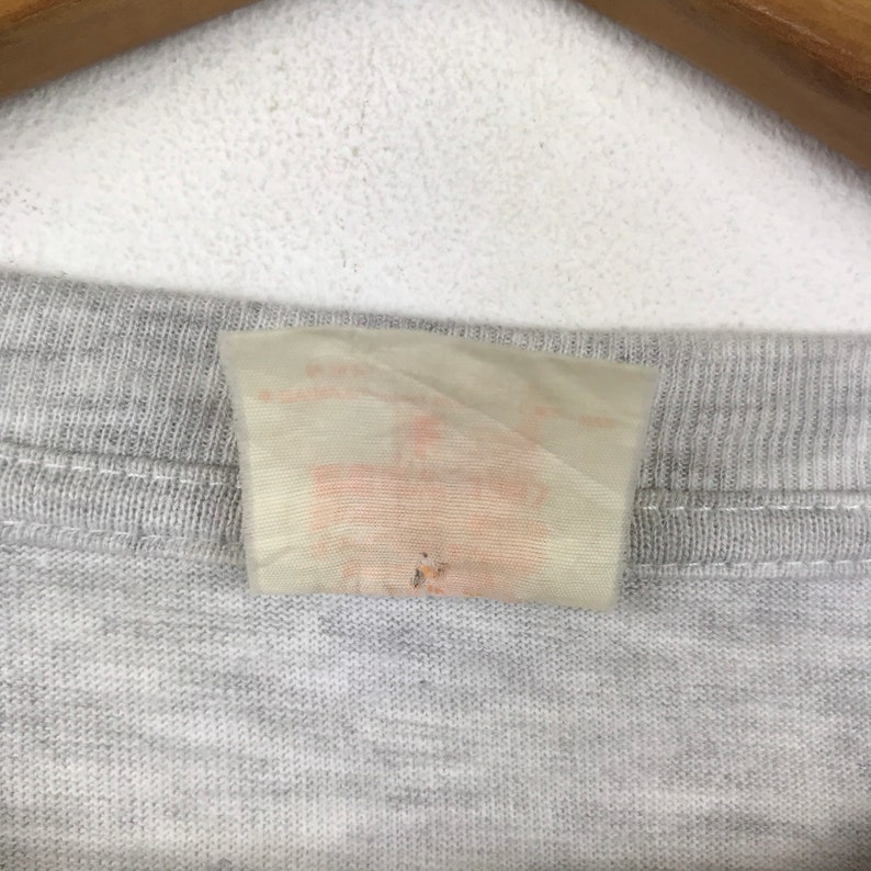 May include: A close-up of a white tag with a faded orange label attached to a gray shirt.