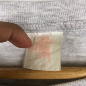 May include: A close-up of a white tag with a pink stain. The tag is attached to a gray t-shirt.