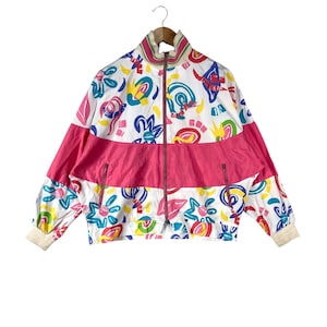 K&ouml;nnte beinhalten: Vintage-Jacke mit Rei&szlig;verschluss und einem lebendigen, abstrakten Muster. Die Jacke hat eine wei&szlig;e Basis mit bunten Formen, einen leuchtend rosa horizontalen Streifen und gerippte B&uuml;ndchen. Der Kragen ist wei&szlig; mit rosa Besatz.