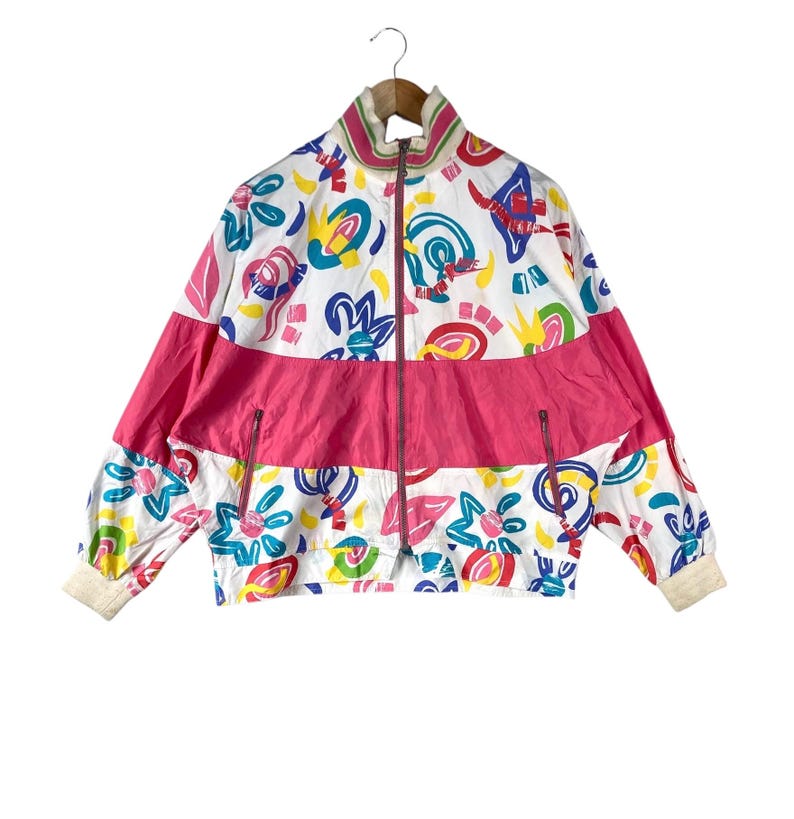 K&ouml;nnte beinhalten: Vintage-Jacke mit Rei&szlig;verschluss und einem lebendigen, abstrakten Muster. Die Jacke hat eine wei&szlig;e Basis mit bunten Formen, einen leuchtend rosa horizontalen Streifen und gerippte B&uuml;ndchen. Der Kragen ist wei&szlig; mit rosa Besatz.