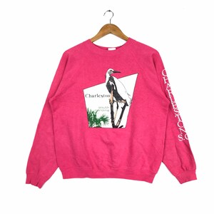May include: Pink sweatshirt with a white egret graphic and the text "Charleston, South Carolina". The left sleeve has the text "Charleston, S.C." printed vertically.