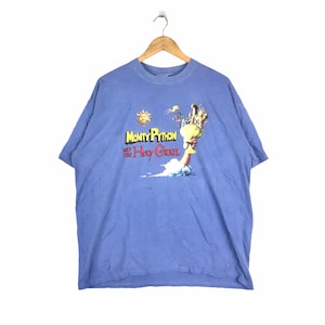 Peut inclure: Un t-shirt bleu clair avec un imprimé graphique de Monty Python et le Sacré Graal. Le graphique représente une main tenant un calice doré avec un soleil et des chevaliers en arrière-plan.