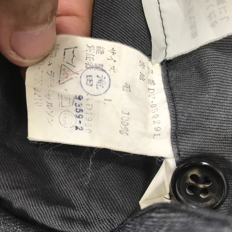 May include: A white tag with black text attached to a dark grey garment. The tag reads "品番D2-05029L 表地 毛 100% I. 縫製泡 責任者田 田D1900 9359-2 サイズ".