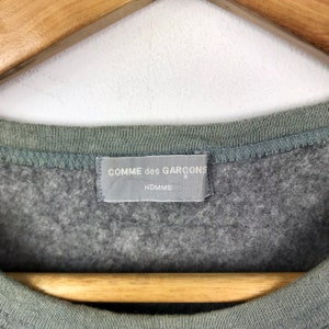Op de afbeelding: Een grijs sweatshirt met een grijs label met de tekst "COMME des GARCONS HOMME"