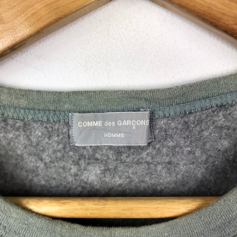 Op de afbeelding: Een grijs sweatshirt met een grijs label met de tekst "COMME des GARCONS HOMME"