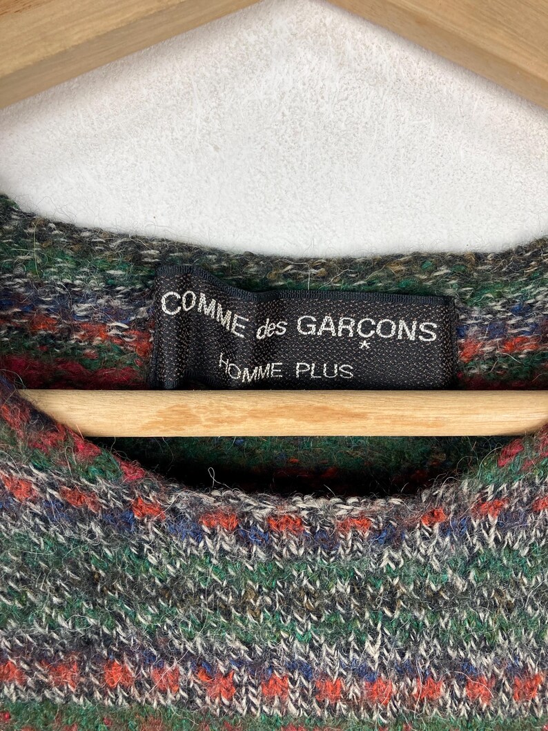 May include: A green, red, and blue patterned sweater with a black label that reads "COMME des GAR&Ccedil;ONS HOMME PLUS".