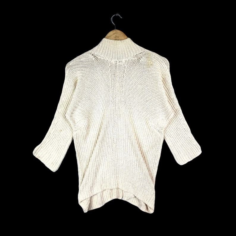 May include: A cream-colored knitted sweater with a mock neck and 3/4 sleeves. The sweater has a relaxed fit and a slightly longer length in the back.