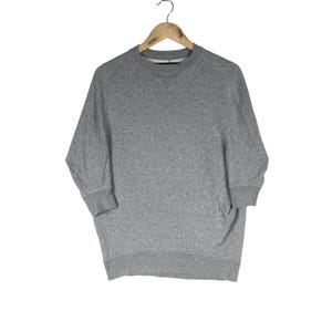 以下が含まれることがあります： 丸首と七分袖のヘザーグレーのスウェットシャツが木製のハンガーに掛けられています。スウェットシャツはリブ編みの袖口とゆったりとしたフィット感です。背景は無地の白です。