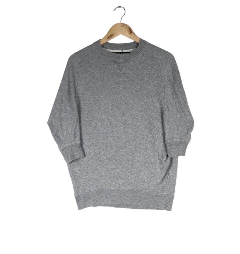 以下が含まれることがあります： 丸首と七分袖のヘザーグレーのスウェットシャツが木製のハンガーに掛けられています。スウェットシャツはリブ編みの袖口とゆったりとしたフィット感です。背景は無地の白です。