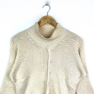 May include: A cream-colored knitted sweater with a mock neck and long sleeves. The sweater has a textured knit pattern.