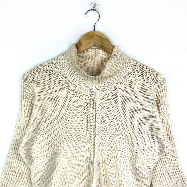 May include: A cream-colored knitted sweater with a mock neck and long sleeves. The sweater has a textured knit pattern.