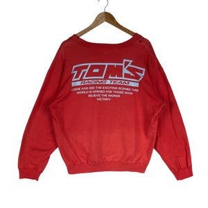 May include: A red long-sleeve sweatshirt featuring the white and light blue text TOM'S RACING TEAM and the phrase COME AND SEE THE EXCITING SCENES THIS WORLD IS OPENED FOR THOSE WHO BELIEVE THE WORDS VICTORY printed on the front chest area.