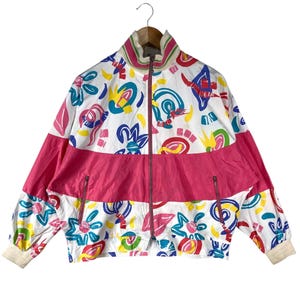 K&ouml;nnte beinhalten: Vintage wei&szlig;e Jacke mit einem lebendigen abstrakten Muster in Blau, Gelb, Rot und Gr&uuml;n. Die Jacke hat einen leuchtend rosa horizontalen Streifen &uuml;ber der Brust und zwei Rei&szlig;verschlusstaschen. Kragen und Manschetten sind cremefarben.