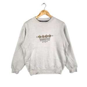 Könnte beinhalten: Ein hellgrauer Pullover mit einem braunen und schwarzen Grafikdruck, der "Refinement and Quality" lautet. Die Grafik enthält auch eine Reihe von braunen Rauten.